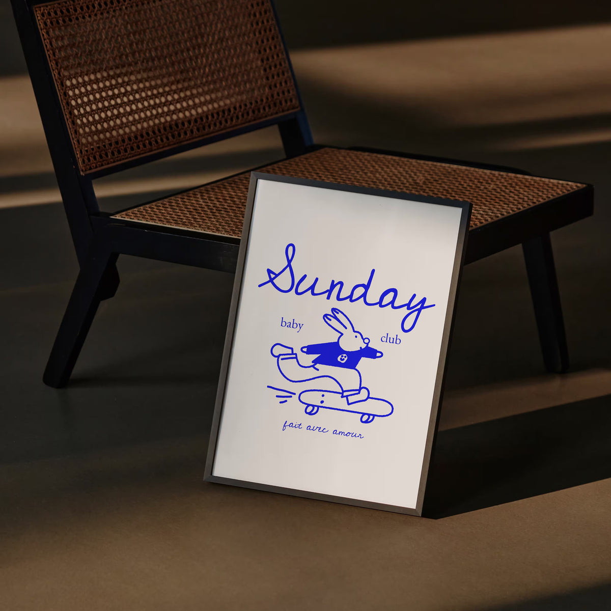 Affiche Sunday Baby Club encadrée posée au sol contre une chaise en rotin – décoration chambre bébé
